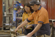 A young apprentice wearing safety glasses and an orange work shirt receives hands-on training from a supervisor in a workshop, illustrating mentorship and skill development in the workplace.